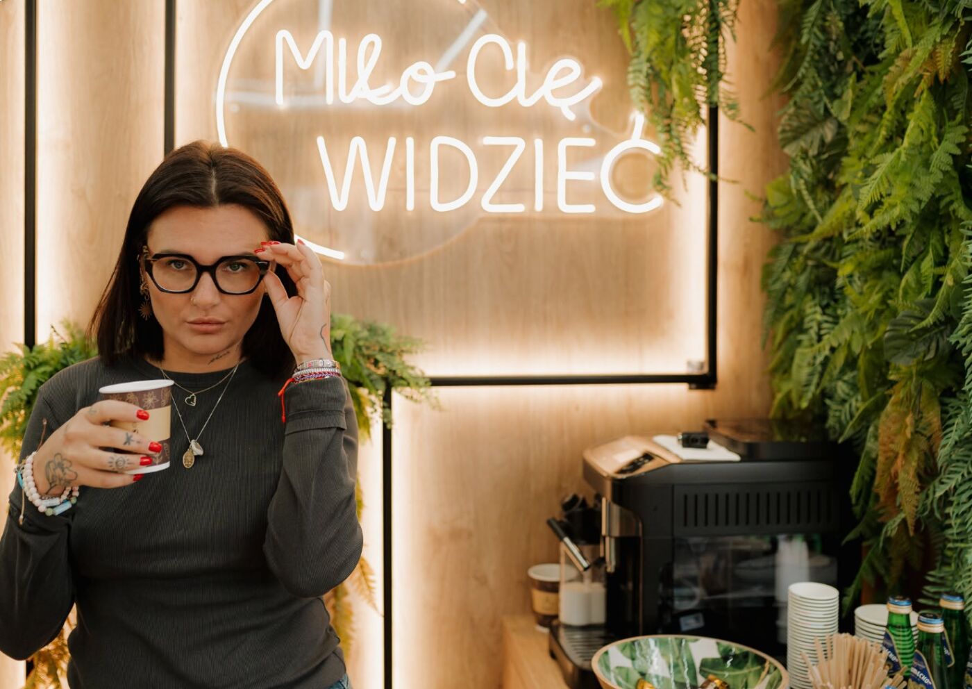 Kobieta poprawia okulary z kubkiem kawy w ręku, w tle neon z napisem Miło Cię widzieć